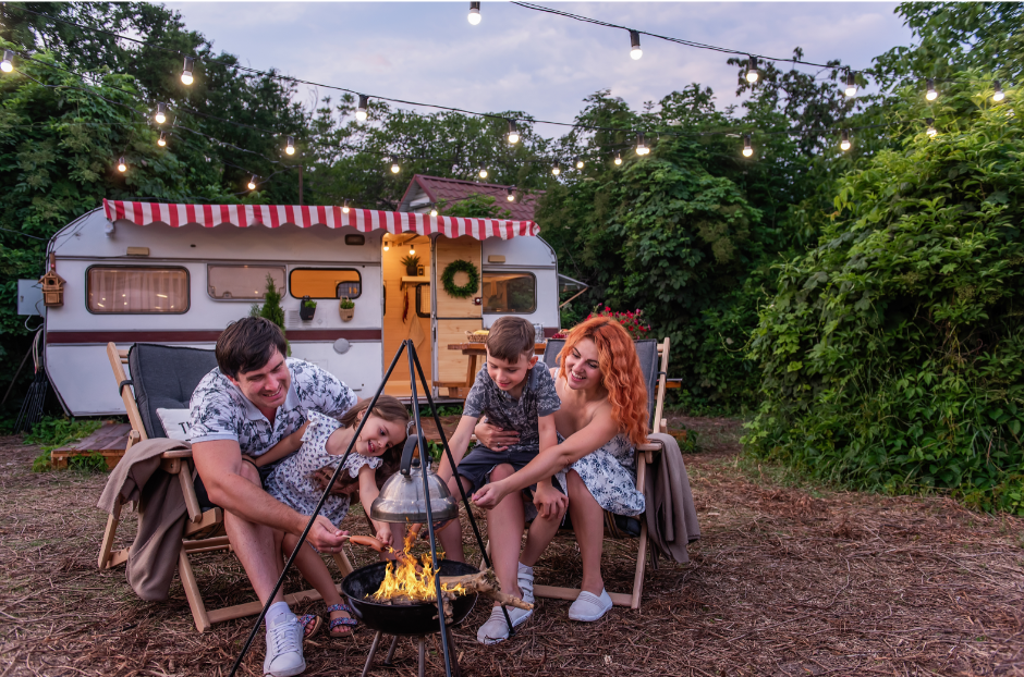 Happy family gathered around a campfire in front of a charming vintage RV decorated with string lights and a red striped awning, representing the guest experience that thrives when an RV park owner runs a financially healthy operation.