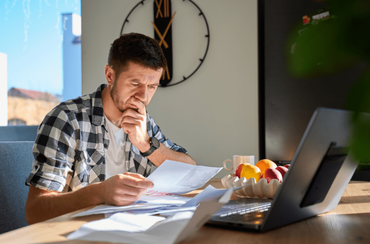 Focused male investor sitting at a home office desk carefully reviewing financial documents and paperwork with a laptop open beside him, representing the due diligence process of evaluating a business or property acquisition