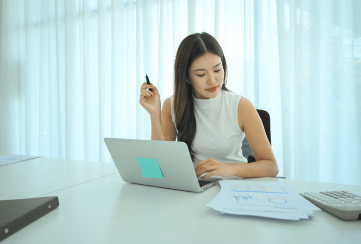 Professional woman sitting at a clean white desk reviewing financial reports on her laptop with a pen in hand, representing the focused financial analysis a fractional CFO brings to your business.