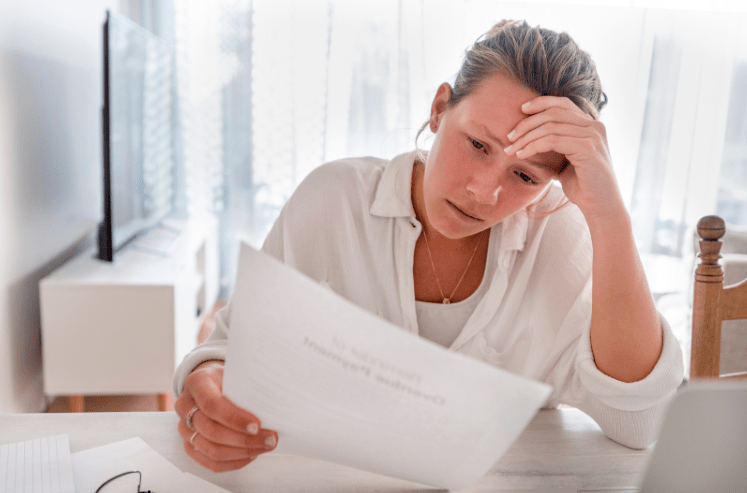 Stressed woman sitting at a home desk with her hand on her forehead reading a financial document with a worried expression, representing the anxiety and overwhelm of discovering a cash flow crisis in your business.