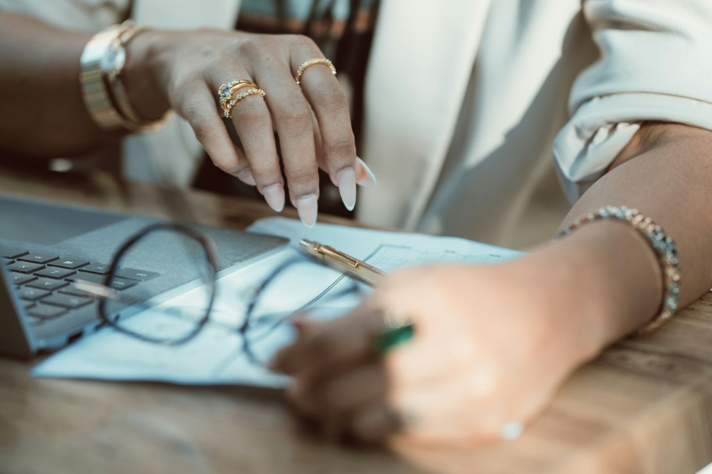 A close up of a woman's hands reviewing financial documents at a desk with a laptop and pen, analyzing the numbers before making a decision