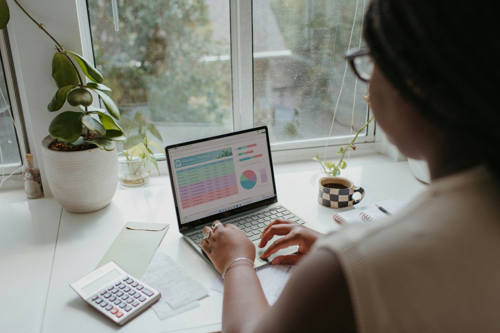 Woman reviewing financial spreadsheet with pie chart on laptop screen, calculator and notebook on desk beside her.
