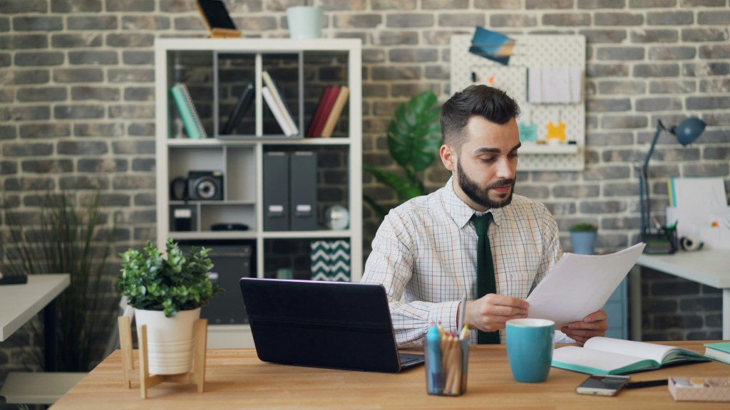 A business professional sitting at a desk reviewing a document with a laptop open, coffee cup nearby, taking a careful look at the details before making a decision.